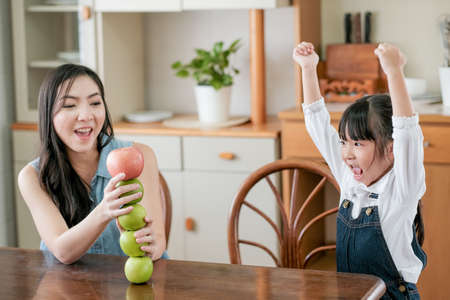 Asian daughter show happy action after play by set apples together on table with her mother in the kitchen. Concept of enjoy with family time of different age of member in their house.の写真素材