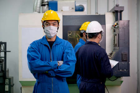 Factory worker man with mask and safety uniform stand with confidence action and stay in front of co-workers work with machine. Concept of good management system for better industrial business.の写真素材