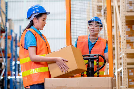 Warehouse woman or factory worker hold package box in front of hand stacker or hydraulic cart that...