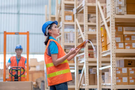 Warehouse woman or factory worker with blue hard hat and uniform stand in front co-worker with cart...