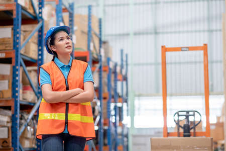 Warehouse woman or factory worker with blue hard hat and uniform stand in front of hydraulic cart in workplace. Concept of good management and happiness of staff during work in business.の写真素材