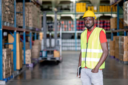 Portrait of African American warehouse man or factory worker with safety uniform smile and stand with confident action in storehouse workplace and stay in front of forklift. He look happy with workingの写真素材