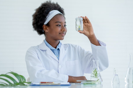 African American young girl hold and look to small glass jar containing piece of plant inside and also smile with happy emotion after finish experiment in laboratory or classroom.の写真素材