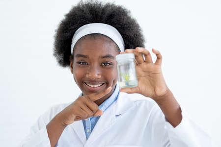 African American young girl point to small glass jar with piece of plant inside and also look to camera and smile with happy emotion after finish experiment in laboratory or classroom.の写真素材