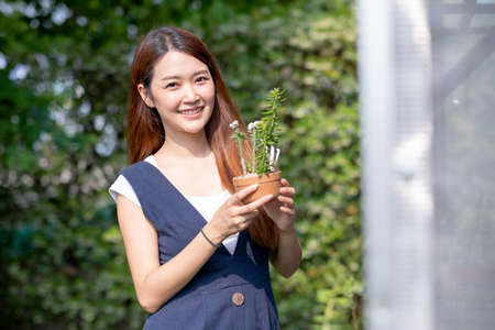Beautiful Asian girl hold pot of plant and stand near green house with day light. Sustainable with grow cactus and other plant concept.の写真素材