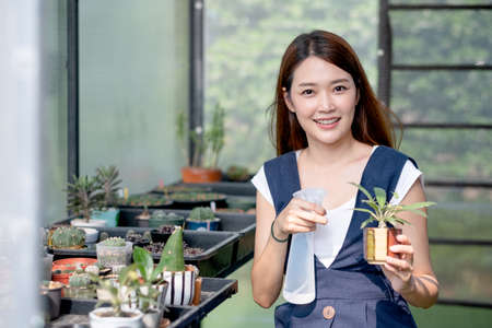 Beautiful Asian girl hold foggy bottle for spraying water to plant in small pot and sit in green garden in her house. Sustainable with grow cactus and other plant concept.の写真素材