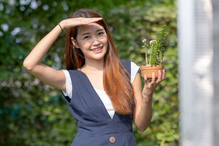 Beautiful Asian girl hold pot of plant with hand shading of light and stand near green house and smiling. Sustainable with small business relate to plant concept.の写真素材