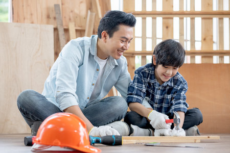 Asian father look carefully son use hammer to hobnailed in their workplace of carpentering with happy emotion. Asian family concept to stay at home and enjoy good relationship hobby together.の写真素材