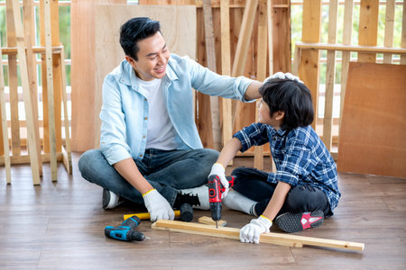 Asian father touch head of his son during drill the timber in home workplace of carpentering with happy emotion. Asian family concept to stay at home and enjoy good relationship hobby together.の写真素材