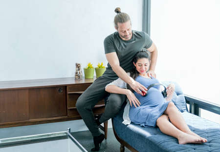 Pregnant woman lie on sofa and hold headphone on her abdomen with husband for baby to listen some...