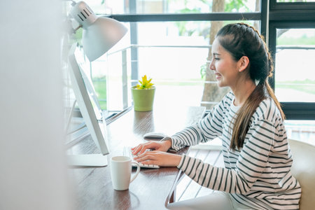 Asian woman look happy by smiling during work with computer in the room with day light. Concept of work at home during pandemic of virus.の写真素材