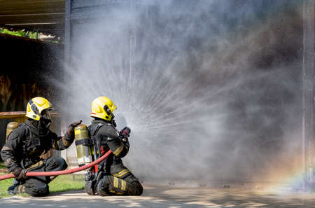Two firefighters sit on floor and spray of water with curtain shape and rainbow reflex occur on water aerosol in front of container containing fire inside.の写真素材