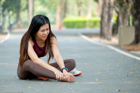 Asian woman sit on road floor and hold her foot look like she got pain and injury during exercise in park or garden in the morning.の写真素材
