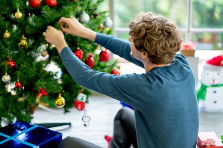 Back of man decorate the Christmas tree in the room of his house and he put many gift box or present around area the he stay.の写真素材