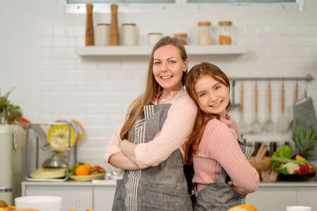 Caucasian woman with her daughter wear apron and lean back stand together also look at camera with smile in home kitchen during cook together.の写真素材