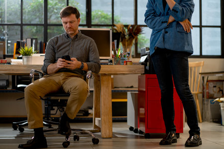 Caucasian business sit on chair and use mobile phone while his co-worker stand beside. He look boring and tired after long time to work.の写真素材
