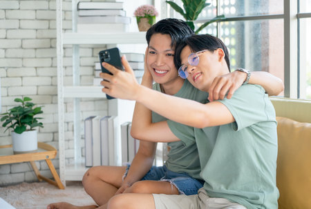Couple gay or lgbt man enjoy with mobile phone together and sitting on sofa near bed in bedroom with day light and they look happy and relaxの写真素材