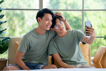 Couple gay or lgbt man enjoy with mobile phone together and sitting on sofa near bed in bedroom with...