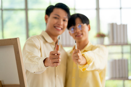 Main focus on paintbrush hold by two gay or lgbt man stand in living room with painting pallet and day light.の写真素材