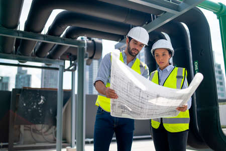 Two professional engineer discuss with blue print of construction site and stay under the floating pipe or pipe hanging on deck of building.の写真素材