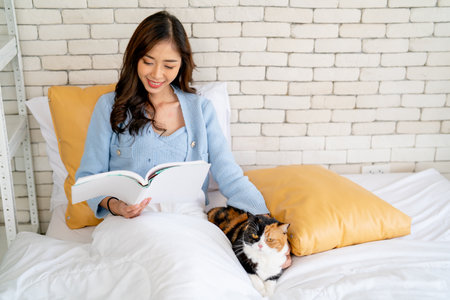 Asian woman sit on bed and relax by read the book also stay and pet  her cat in bedroom in concept of good relationship and bonding between owner and pet.の写真素材