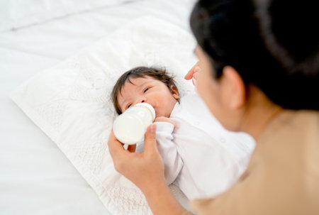 Back of Young Asian mother give milk in bottle to newborn baby lie on bed and take care with...