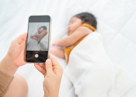 Close up hands of mother use mobile phone to take photo of her newborn baby sleep on bed with day light.の写真素材