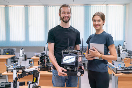 Two young Caucasian professional technician or engineer workers stand with one hold small robitic machine and the other one hold tablet stand in factory room workplace and look at camera with smiling.の写真素材
