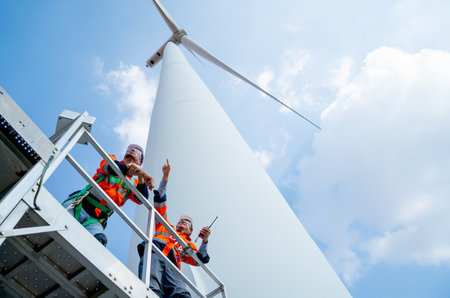Lower view of two engineers or technician workers stand on base of big windmill or wind turbine and point or look to left side with blue sky.の写真素材