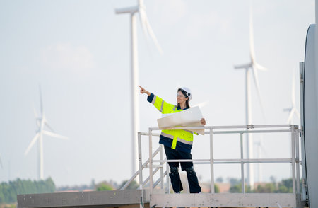 Pretty engineer or technician woman worker hold drawing paper and stand on base of windmill or wind turbine and point to left side and stay in front of windmill cluster in background with blue sky.の写真素材