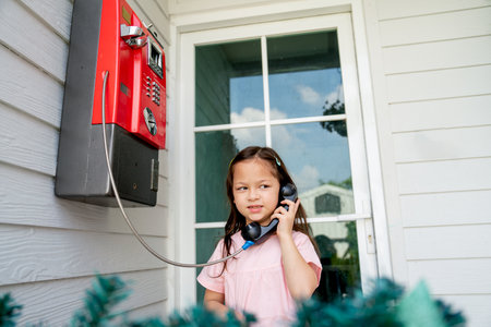 Cute little Asian girl enjoy to play with payphone in front of door of one room in theme park with...