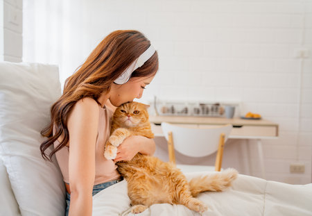 Pretty Asian woman with headphone hold and kiss orange cat sit on the bed in bedroom of her house.の写真素材