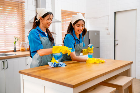 Two Asian housekeeper or housemaid women enjoy to work and teasing each other with one woman spray cleaner to top surface during clean table in kitchen at customer house.の写真素材