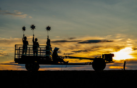Silhouette of children hold small windmill and stand on E-taen farm tractor with driver to go back home in early morning with warm sunrise light.の写真素材
