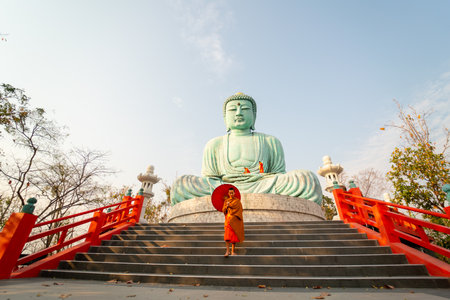 Wide shot of young Asian monk hold red umbrella and walk down on stair in front of green big buddha statue with soft light in the morning.の写真素材