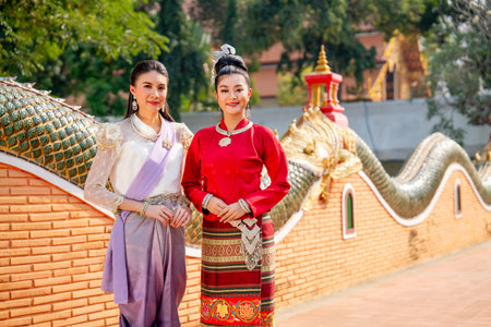 Portrait of two Asian women with Thai traditional dress stand together in the way to ancient pagoda with beautiful art and architecture, they also smile and look at camera.の写真素材