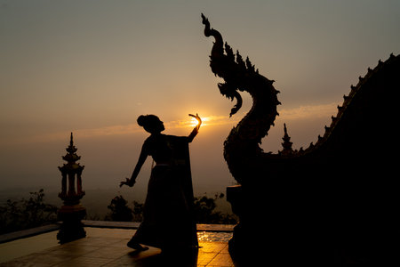 Silhouette of woman wear Thai traditional dress and stand with Thai dance in front of naga sculpture in temple with sunrise in background.の写真素材