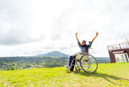Wide shot of Asian senior man sit on wheelchair and action of raise his hands up like stretching also stay near cliff with mountain and grass field in the background.の写真素材