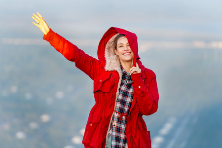 Beautiful woman stand near cliff with hold coat hood and wave hand on the mountain with warm light of sun rise and she look happiness with smiling also look at camera.の写真素材