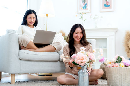 Pretty Asian woman sit on sofa and use laptop to work while young woman sit on the floor and enjoy to flower arrangement in living room.の写真素材