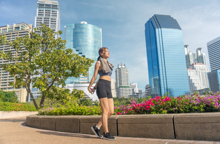 Wide shot of Asian sport woman walk and  exercise in public garden with high building of big city in...
