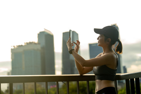 Asian sport woman use mobile phone to take photo and relax after exercise in public garden with high building of big city in background and soft warm light.の写真素材