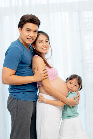 Asian family with father, pregnant mother and little girl hug together and stand in front of white...
