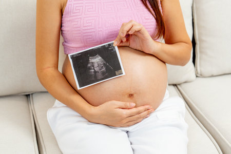 Close up pregnant woman hold with left hand ultrasound image of her baby and sit on sofa in living...