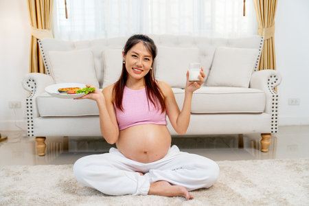 Asian pregnant mother sit on the floor of living room with holding disk of food and milk and look at camera with smiling.の写真素材