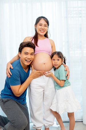 Vertical image of Asian father and little daughter hug and touch to belly of pregnant mother and express happiness with smiling in front of white curtain.の写真素材