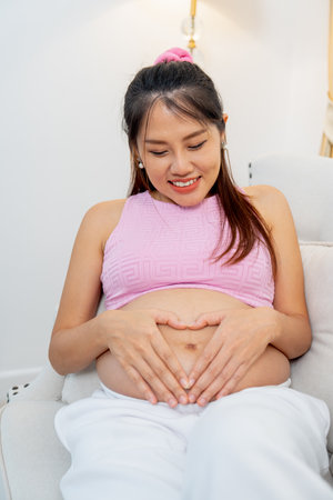 Vertical image of Asian pregnant woman sit on sofa and put hands on her belly with heart shape and smiling.の写真素材