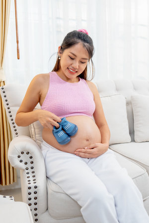 Vertical image of Asian pregnant woman sit on sofa and hold pair of blue shoes and put to touch her...