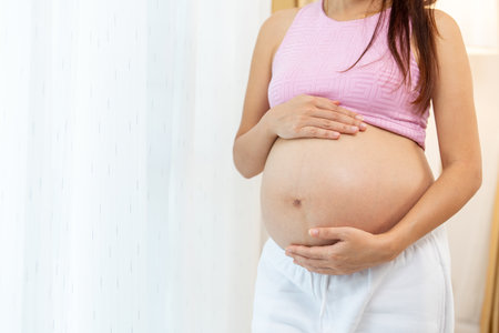 Close up pregnant woman stand with touch her belly in front of white curtain look relax with happiness.の写真素材
