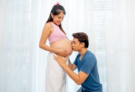 Asian man sit down and kiss belly of pregnant woman to express love and care in front of white curtain in their house.の写真素材